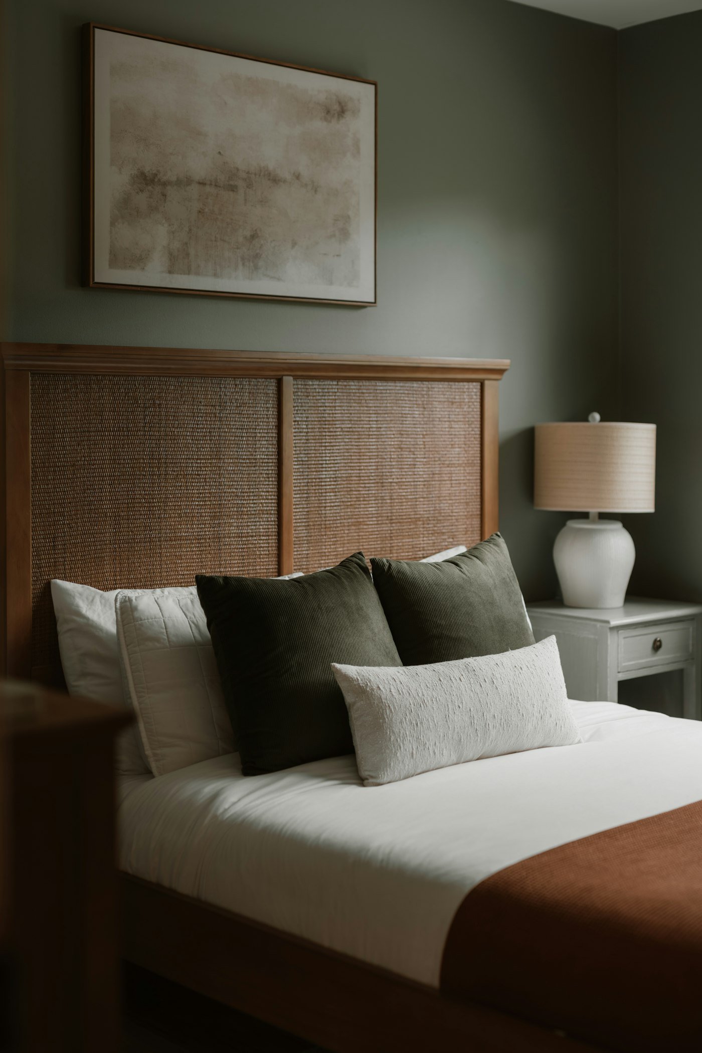 Chambre au mur vert de gris avec tête de lit cannée, lampe en céramique et coussins kaki